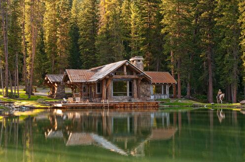 Обои на рабочий стол Деревянный дом с металлической крышей на берегу озера, рядом с домом деревянная терраса с креслами, человек на лошади на берегу, на фоне густого леса из хвойных деревьев