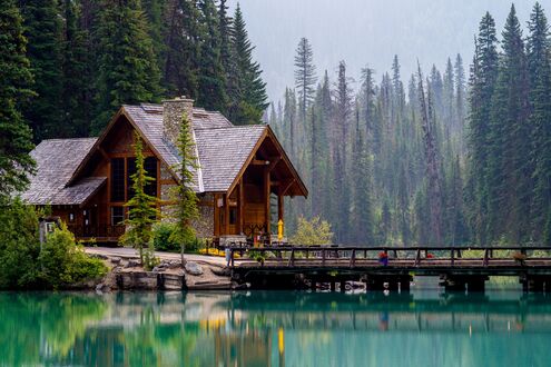 Обои на рабочий стол Эмералд Лейк Лодж / Emerald Lake Lodge, Канада / Canada, деревянный дом с каменным дымоходом и деревянный мост через бирюзовое озеро, на фоне густого хвойного леса и туманного неба