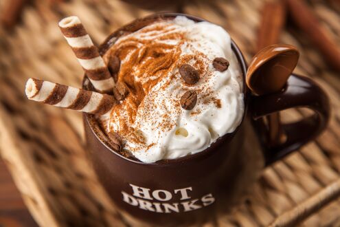 Обои на рабочий стол Коричневая кружка с надписью (HOT DRINKS) со взбитыми сливками, корицей, кофейными зернами, вафельными трубочками и шоколадной ложкой, на плетеной подставке на размытом фоне с палочками корицы
