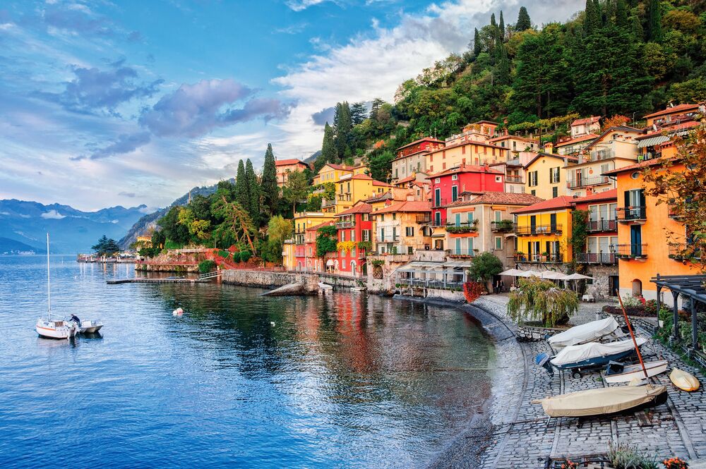 Обои для рабочего стола Варенна / Varenna, Италия / Italy, лодки на синей воде и на мощеном берегу, на фоне разноцветные дома на зеленом склоне холма с деревьями, горами и облачным небом