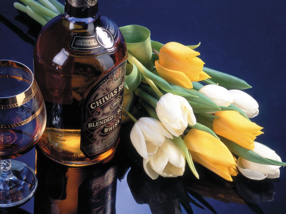 Обои для рабочего стола Бутылка виски Chivas Regal и бокал с напитком на переднем плане, рядом букет из желтых и белых тюльпанов на темном фоне