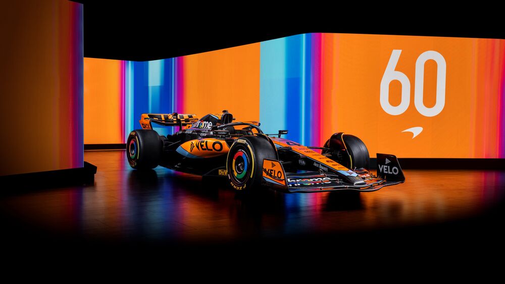 Обои для рабочего стола McLaren F1 гоночный автомобиль черного и оранжевого цвета на темном полу, (VELO, chrome) на фоне светящихся панелей с оранжевыми и синими полосами и числом (60)
