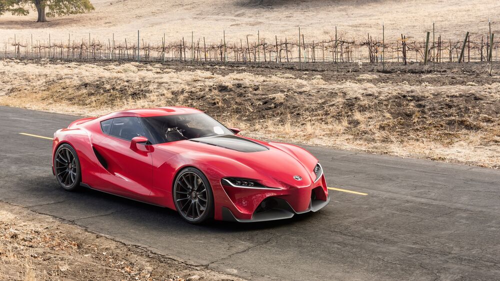 Обои для рабочего стола Красный спортивный автомобиль Toyota Supra едет по асфальтированной дороге с желтой разметкой, на фоне сухих холмов с виноградником и редкими деревьями