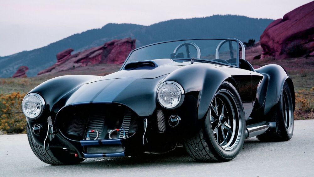 Обои для рабочего стола Черный автомобиль Ac Cobra / Shelbi с синими полосами припаркован на дороге, на фоне гор с красными скалами и редкой растительностью под пасмурным небом