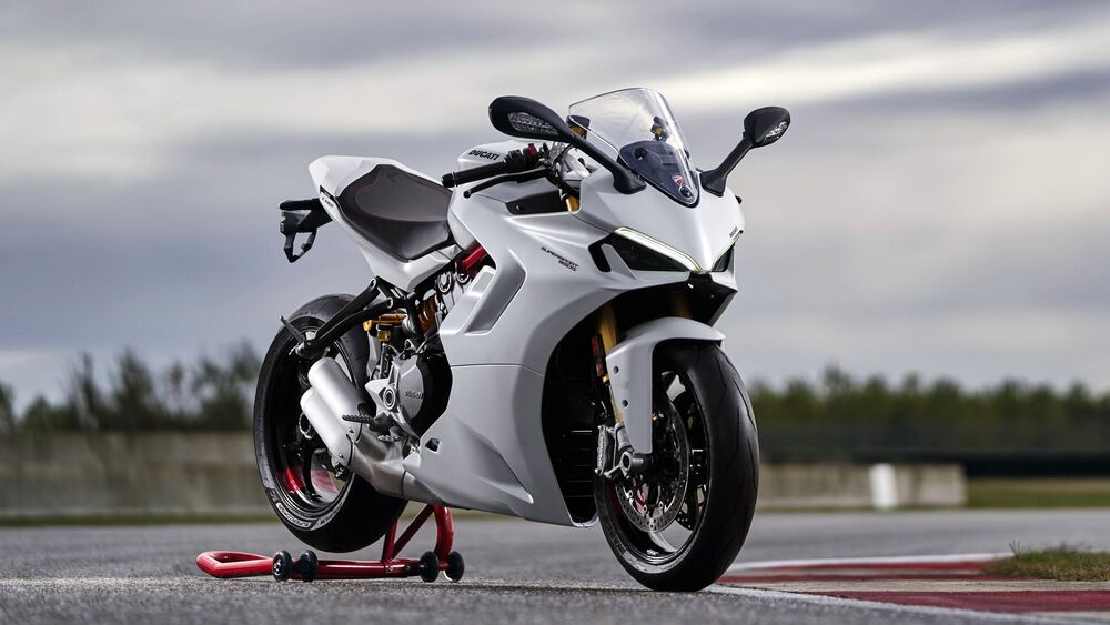 Обои для рабочего стола Белый мотоцикл Ducati / Дукати SuperSport / СуперСпорт 950 / 950 S / С на красной подставке на асфальте гоночной трассы на фоне размытых деревьев и пасмурного неба