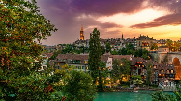 Обои на рабочий стол Берн / Bern, Швейцария / Switzerland, зеленые деревья и бирюзовая река Ааре, исторические здания с освещенными окнами на берегу реки, на фоне башни собора и заката с облаками