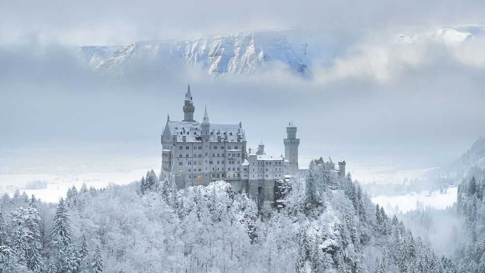 Обои для рабочего стола Замок Нойшванштайн / Neuschwanstein Castle, Германия / Germany, на заснеженном холме среди деревьев, на фоне заснеженных гор и облаков