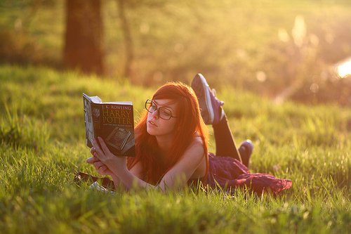 Фото Рыжеволосая девушка в очках  лежа на поляне читает книгу