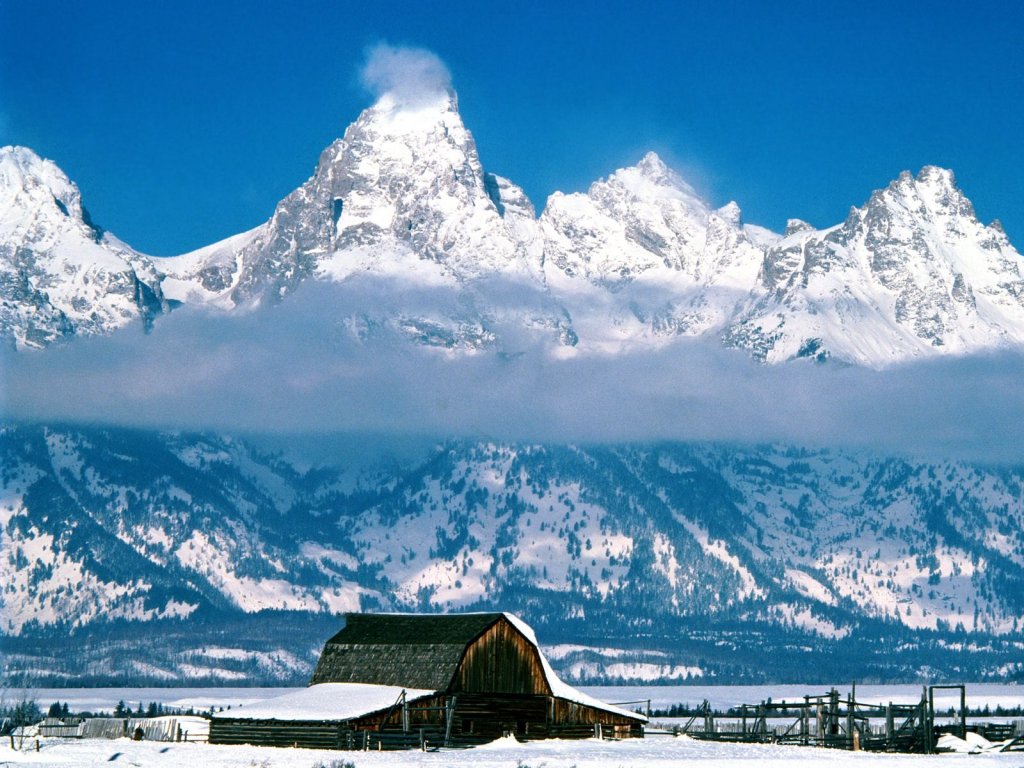 Фото Заснеженные пики гор, расположенные выше облаков, на переднем плане деревянное сооружение для зимовки домашнего скота