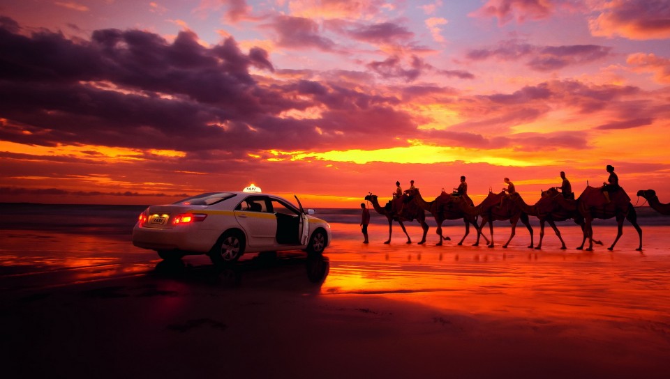 Фото Автомобиль-такси (TAXI) остановился, уступая дорогу каравану верблюдов с погонщиками на фоне багряного неба,