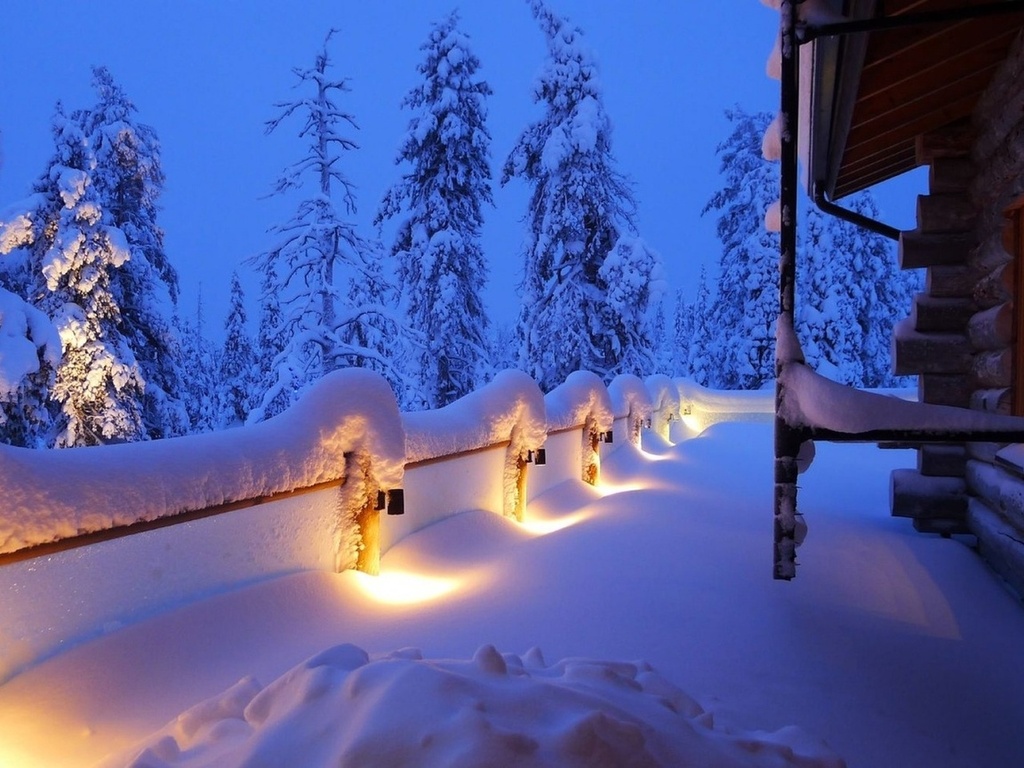  Деревянный забор с горящими фонарями, покрытый густым слоем снега, домик из рубленных бревен на фоне ночного неба