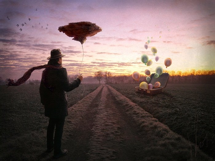 Фото Мужчина стоит в поле, держа на веревочке парящий остров и смотрит на воздушные шары летящие из чемодана