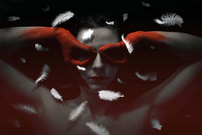 Фото Девушка с раскрашенными в красный цвет руками в виде перчаток, держит руки у глаз, вокруг летают перья, сюрреалистические работа фотохудожницы Валерии Беловой / Valeriya Belova