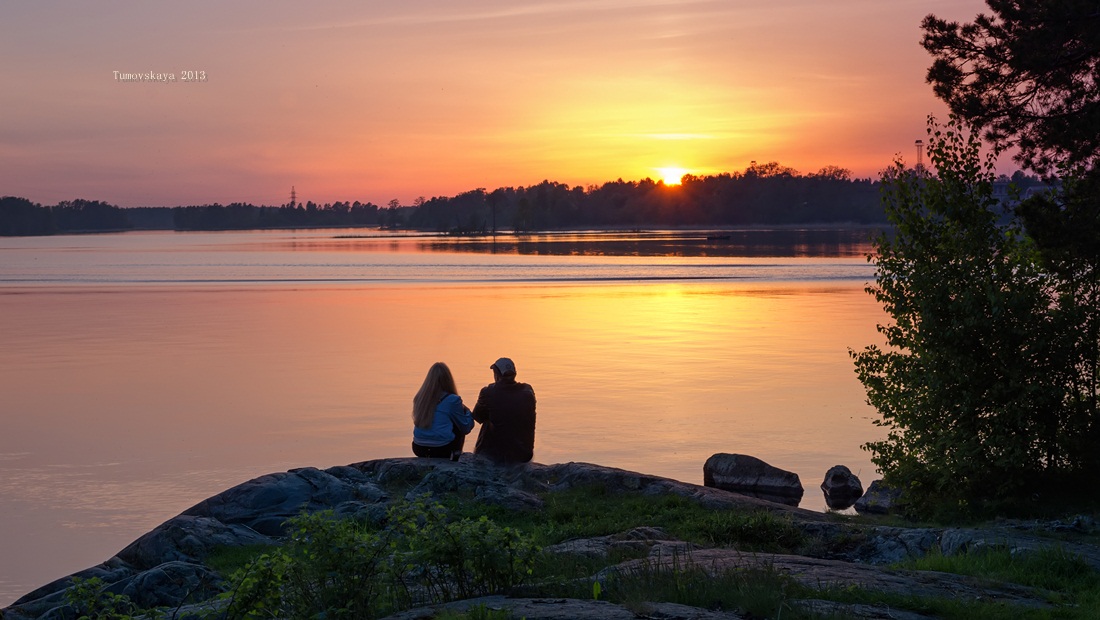 Фото Супружеская пара, сидящая на каменистом берегу озера на фоне заходящего за верхушки деревьев солнца, фотография Екатерины Тумановской