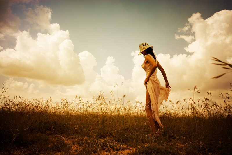 Фото Девушка в белой шляпе и длинном платье стоит в поле на фоне облачного неба