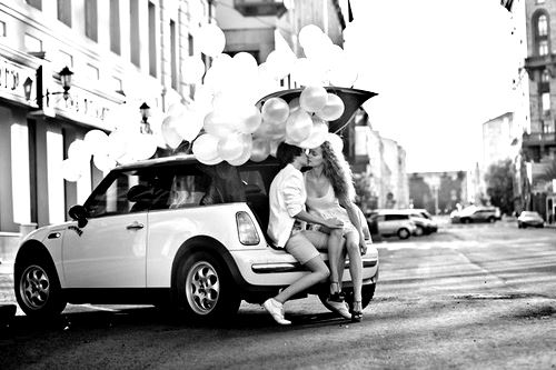 Фото Девушка с мужчиной, сидя, целуются в багажнике белой машины, на которой много шаров, посреди города