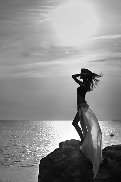Девушка стоит у моря на скале, взявшись за волосы на фоне солнца, моря и неба