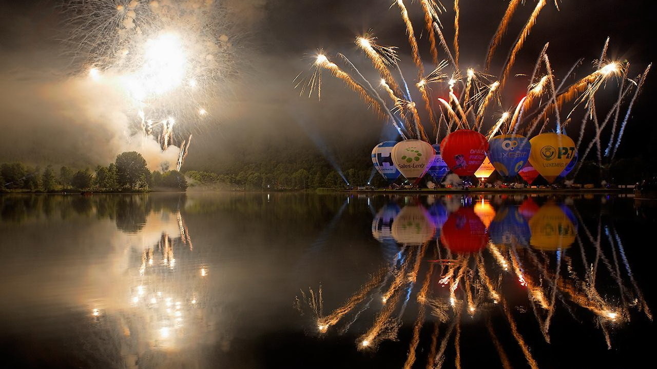 Фото Разноцветные воздушные шары, находящиеся на берегу озера, готовятся к старту в ночное небо на фоне огненных вспышек фейерверка