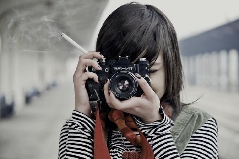 Фото Девушка в полосатой блузке и с сигаретой в руке, смотрит в видоискатель фотоаппарата Зенит TTL / Zenit TTL