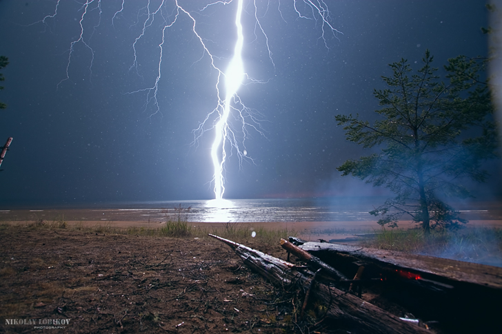 Фото Сильный разряд молнии над морем, попал в лежащее на берегу сухое дерево, вызвал его возгорание и появление легкого, синего дыма, автор NIKOLAY LOBKOV