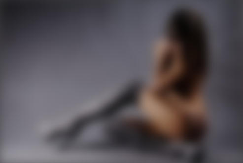 Фото Обнаженная модель Юрис Мария, сидящая полубоком опустив голову, с вытянутой одной ногой вперед, в серых сапогах ботфортах, на сером фоне