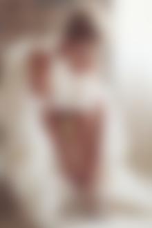 Фото Девушка со светло-русыми волосами в свадебном платье наклонилась вперед к обнаженной ноге, чтобы застегнуть застежку на босоножке, на фоне размытой стены, автор Соня Хегай