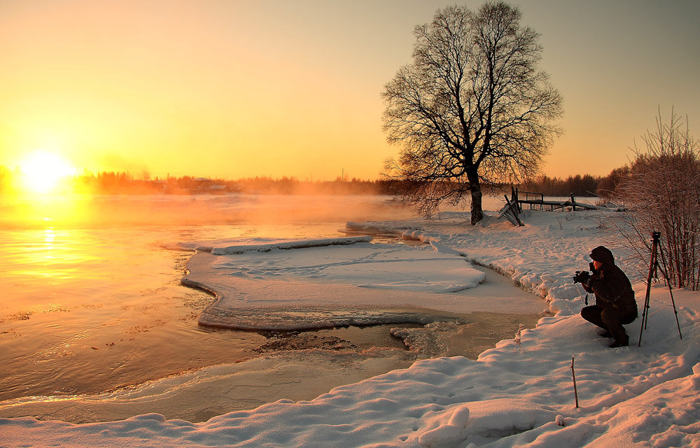 Фото Мужчина-фотограф, стоящий на заснеженном берегу водоема, покрытого у берега кромкой льда, пытается сфотографировать восходящий диск ослепительного утреннего солнца, автор Александр Коротков