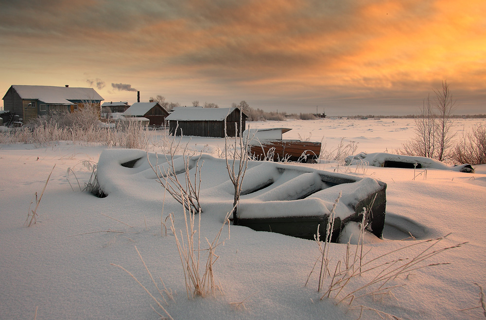 Фото Деревянная лодка, занесенная снегом, находящаяся на берегу замерзшего водоема с расположенными рядом деревянными домами на фоне заката, автор Александр Коротков