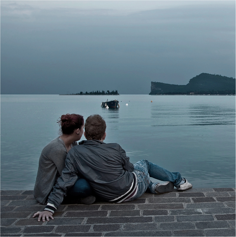 Фото Парень и девушка, тесно прижавшись друг к другу, сидящие на брусчатой морской набережной, наблюдают за лодкой, стоящей в воде напротив их на фоне пасмурного неба на вечернем небосклоне, автор Сергей Гаспарян