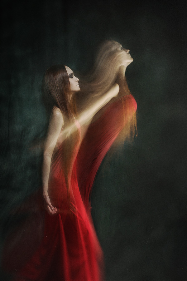 Фото Девушка с длинными каштановыми волосами стоит в профиль в красном до пят платье на темном фоне, и ее душа устремляется вон из тела