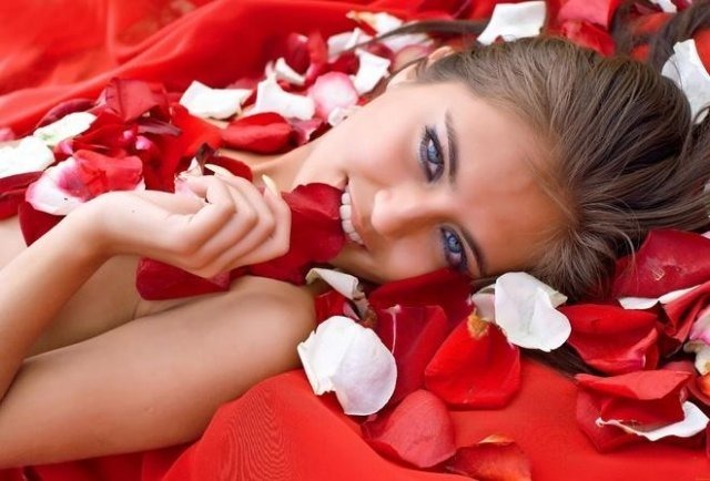 Фото Девушка с голубыми глазами лежит на красной ткани, среди лепестков цветов и держит в зубах лепесток