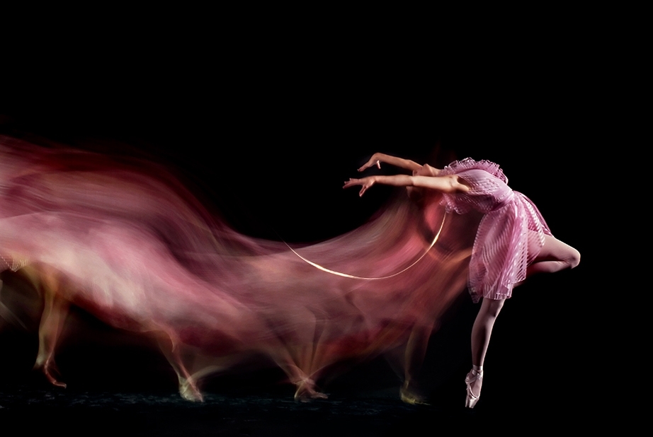 Фото Девушка балерина на черном фоне в розовой одежде и пуантах на ногах делает танцевальное движение, оставляя за собой следы размытости ее движения