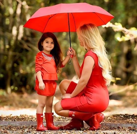 Фото Мама и дочка в красных платьях держат руками зонт находятся на дорожке усеянной осенними листьями