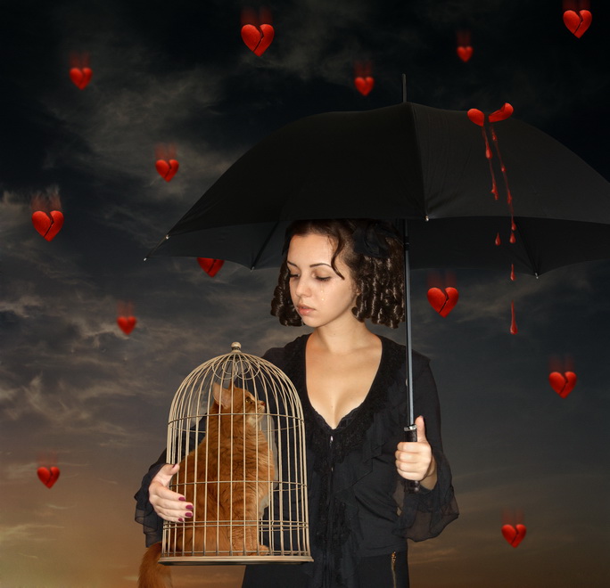 Фото Девушка под зонтом держит птичью клетку с котом, укрываясь от дождя из красных сердечек