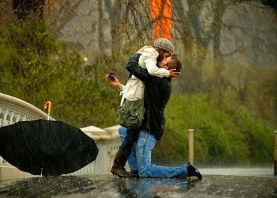 Фото В дождь, откинув зонт в сторону, опустившись на колени, мужчина делает предложение руки и сердца, вместе с обручальным кольцом, своей возлюбленной. Девушка целует парня в ответ