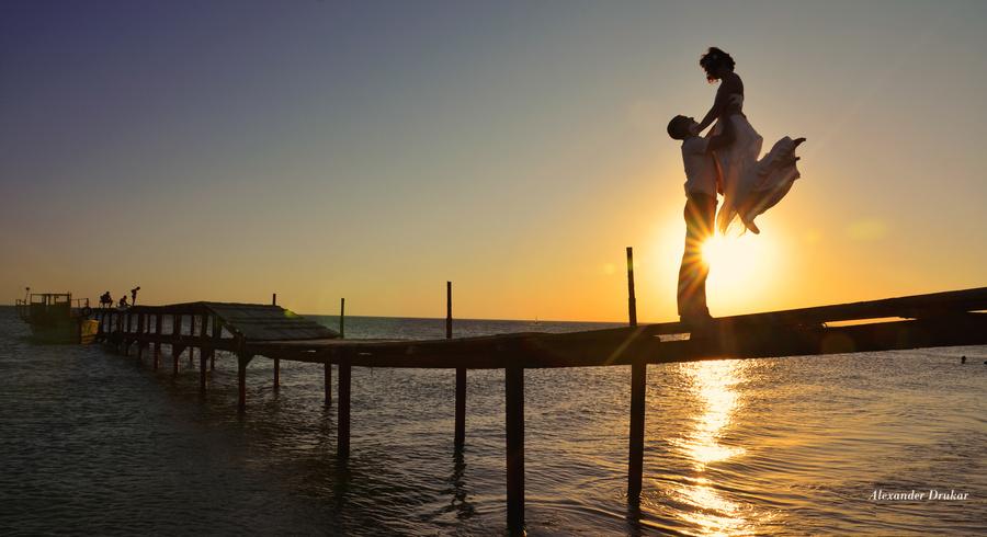 Фото Парень поднял девушку над собой, взяв ее за талию. Оба стоят на морском причале. Вдали садится Солнце. Фотограф Александр Друкар