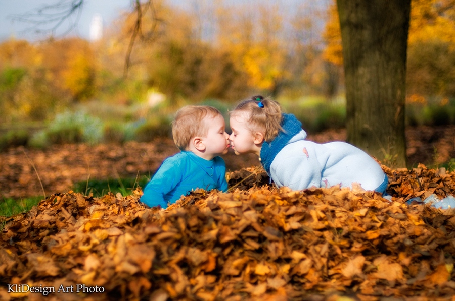 Фото Маленькая девочка целует маленького мальчика, сидя в сухой листве (KliDesign Art Photo)