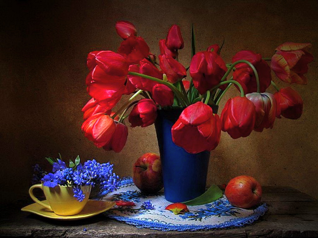 Фото На салфетке стоит синяя ваза с красными тюльпанами, рядом стоит блюдце с чашкой, в которой лежат веточки сирени и возле вазы лежат яблоки
