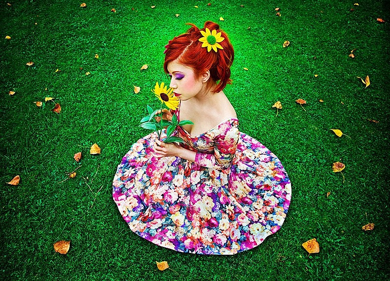 Фото Девушка с желтым цветком в волосах, присела на зеленой лужайке расправив платье кругом и держит в руках желтый цветок