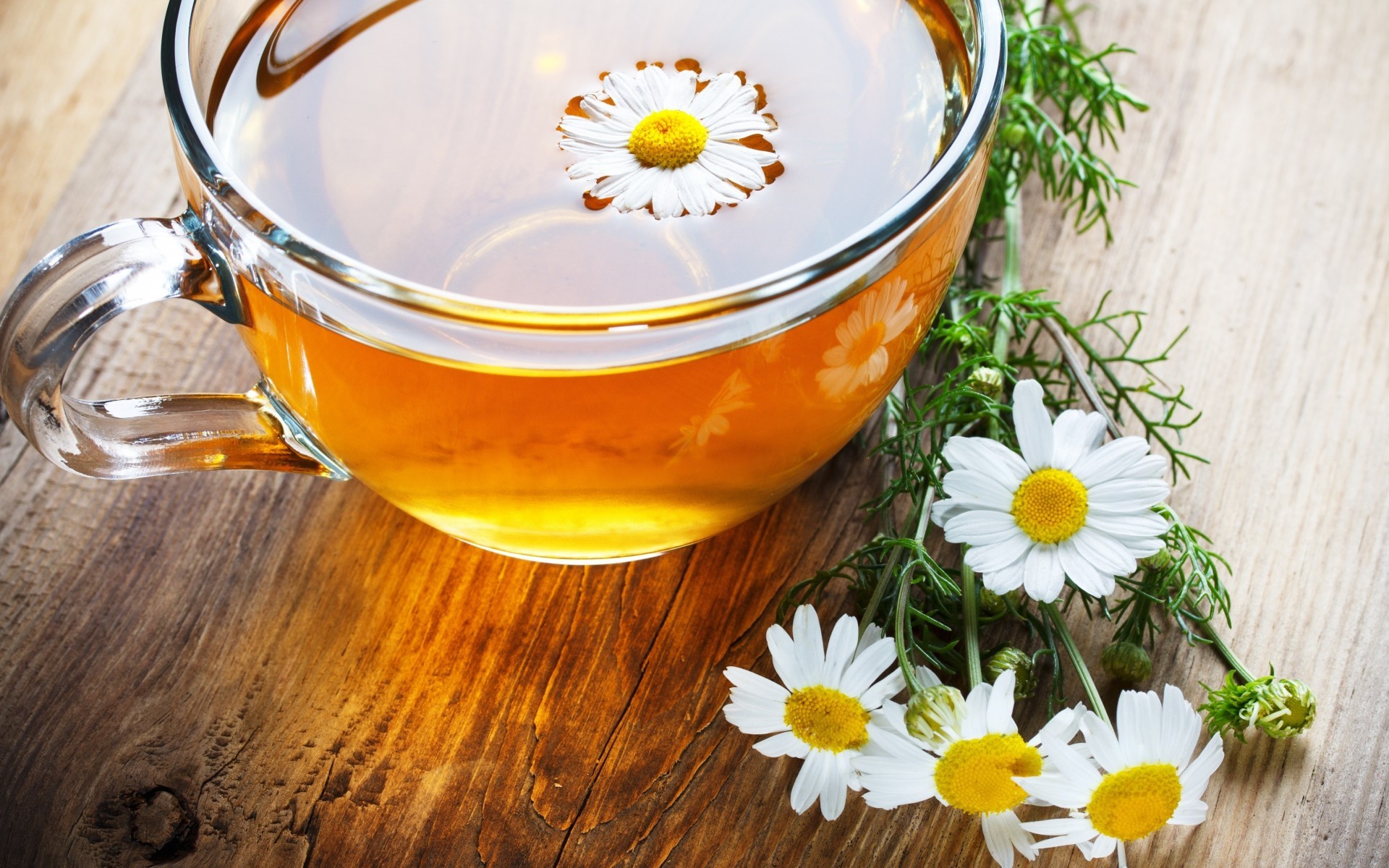 Фото Чашка из прозрачного стекла с чаем и цветком ромашки в чае стоит на столе. Рядом лежит букет из четырех ромашек