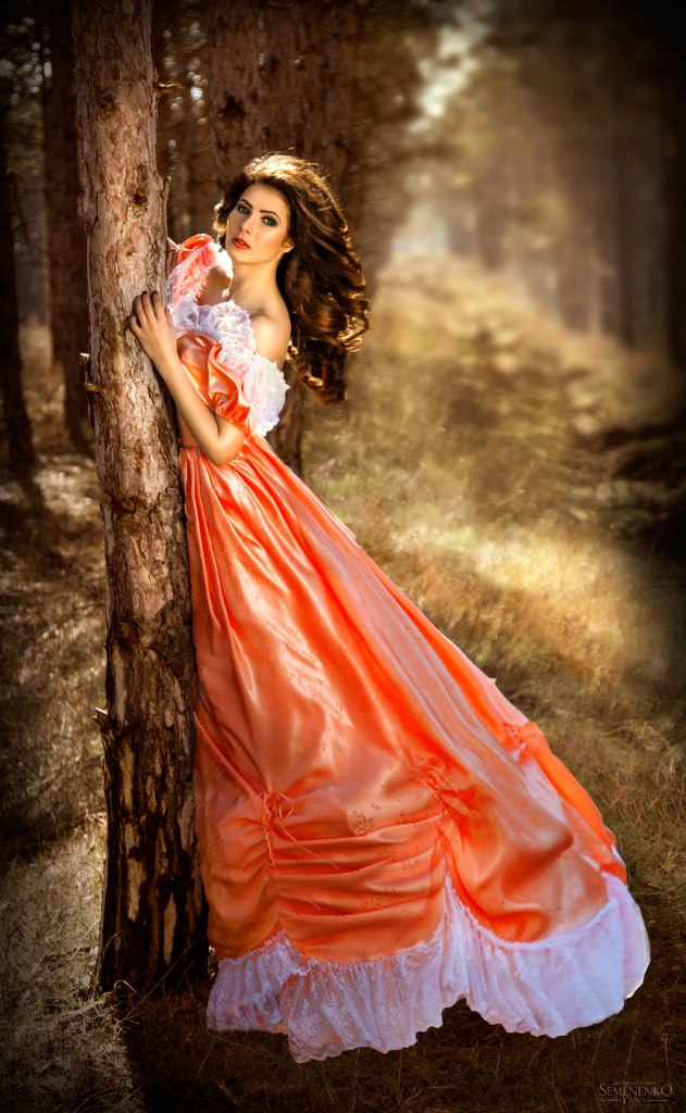Фото Девушка шатенка с длинными волосами в ярком оранжевом платье с белыми воланами стоит у дерева в лесу, фотограф Яна Семененко, модель Диана Сипливец