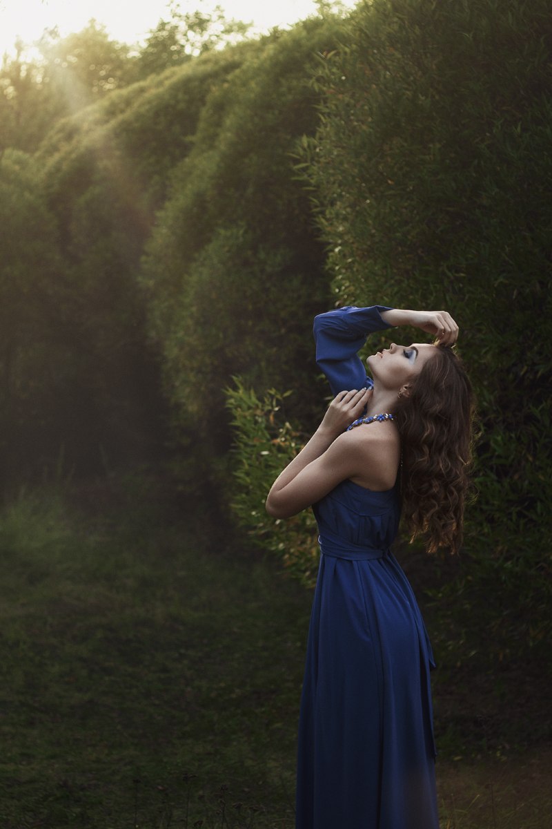 Фото Девушка - шатенка с длинными волосами в синем платье с открытым левым плечом стоит, наклонив голову назад, среди зеленых деревьев, освещенная солнцем, фотограф Иван Копченов, модель Алена Сарсарова