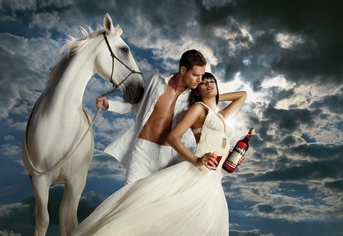 Фото Актриса Ева Мендес / Eva Mendes, в белом платье, держит в руке стакан с красным, горьким ликером, ее обнимает мужчина, одной рукой он держит коня, в другой бутылку Campari, пара стоит на фоне облаков