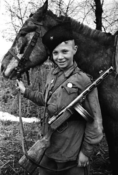 Фото Мальчик в кубанке со звездой, с автоматом и орденом на груди держит лошадь под уздцы, сын полка