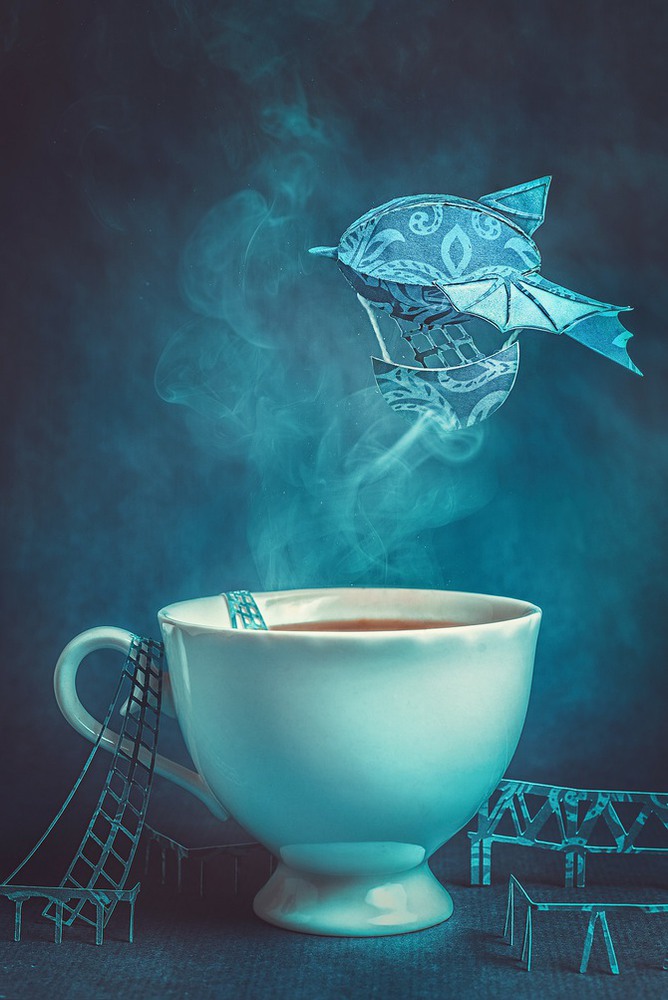 Фото Бумажный дирижабль, проплывающий над чашкой с дымящимся кофе, вокруг которой построены бумажные мосты, фотограф Dina Belenko / Дина Беленко
