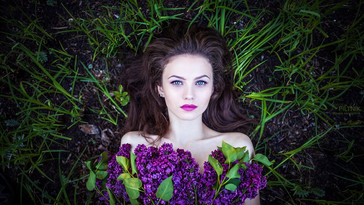 Фото Девушка с темными длинными волосами, голубыми глазами лежит на земле среди травы с букетом сирени, фотограф Сергей Пилтник