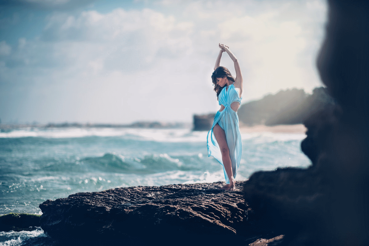 Фото Красивая девушка стоит на берегу моря в прозрачном голубом платье  подняла руки над головой