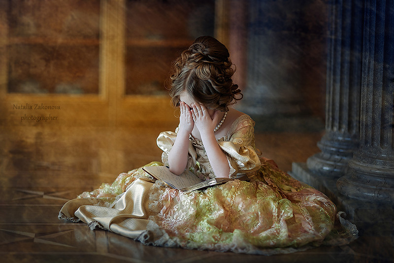 Фото Маленькая девочка грустная c высокой прической, в пышном платье сидит на полу держа на коленях книгу, закрыла руками лицо, на фоне старинного помещения. Фотограф Наталья Законова / Natalia Zakonova