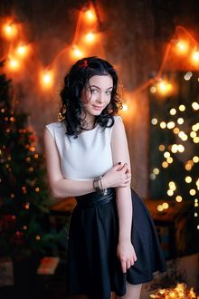 Фото Темноволосая милая девушка в белой кофточке и черной юбке, с тату на руке стоит на фоне праздничного новогоднего фона. Фотограф Лилия Ульянова