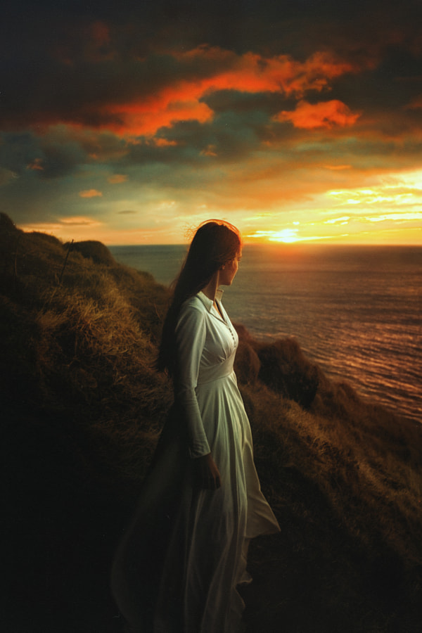 Фото Девушка в длинном белом платье стоит на обрыве у моря, на фоне заката, фотограф TJ Drysdale